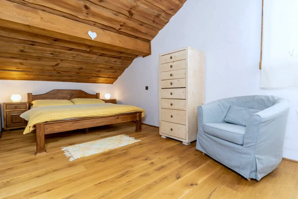 Spálňa pod šikminou: manželská posteľ so žltou posteľnou bielizňou, bledomodré kreslo a vysoká komoda.