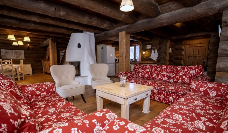 Obývačka, krb, jedálenský stôl a kuchyňa v jednom priestore, v popredí červené kvetované sedačky.