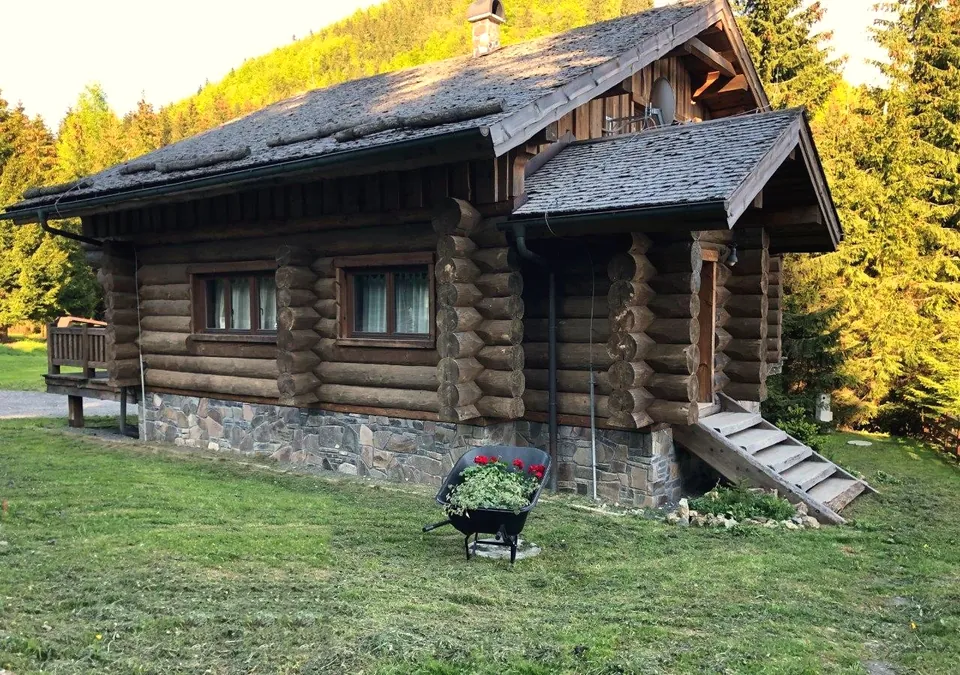 Zrub v lete: rohy z guľatiny, schody ku vchodu, kamenná podmurovka a fúrik s červenými kvetmi na zelenom svahu.
